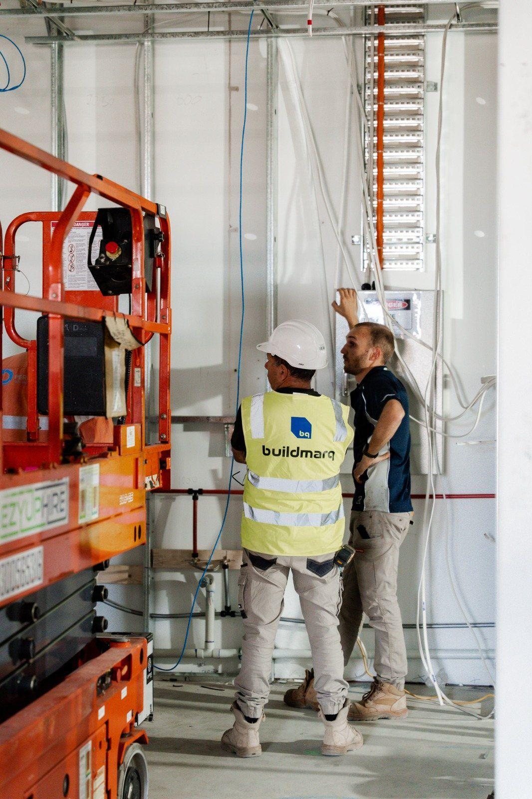 Two people talking beside an electrical board on a fit-out site, one wearing a Buildmarq hi-vis vest, with an orange scissor lift on the left.