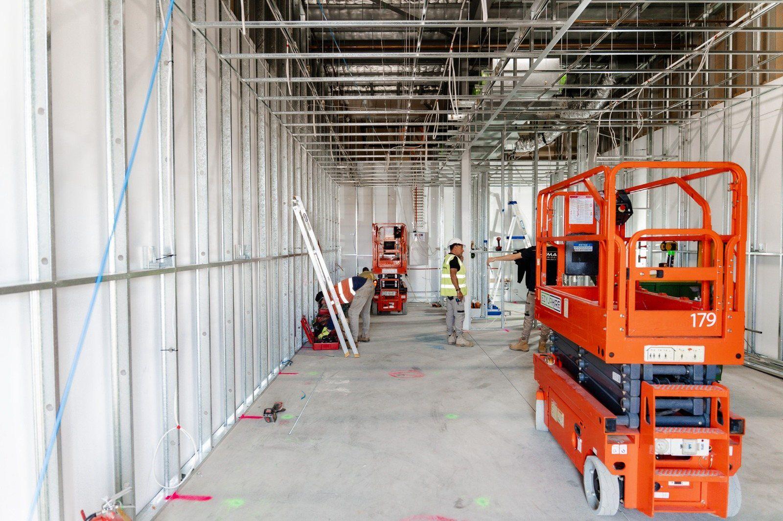A long fit-out corridor with exposed steel framing and services overhead, an orange scissor lift on the right and people working further along.